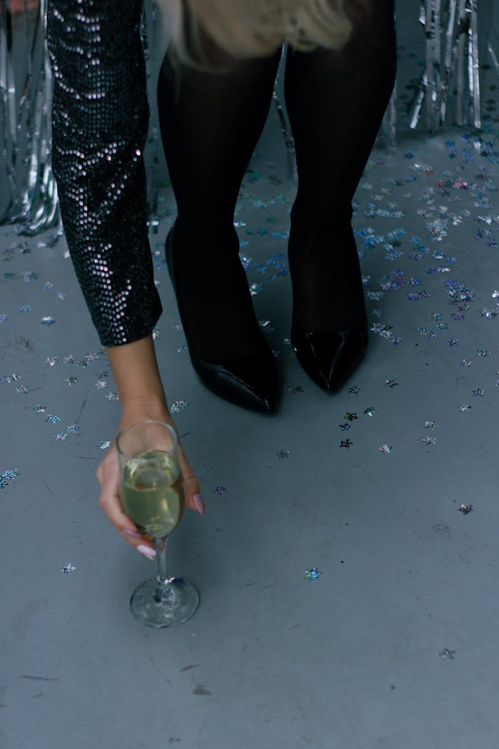 Close-up of a woman in black heels picking up a champagne glass amid party confetti.